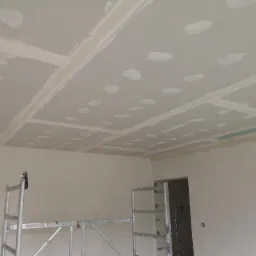 Sufit z płyt kartonowo-gipsowych w trakcie szpachlowania, widoczne łączenia i miejsca po wkrętach, metalowe rusztowanie w dolnej części kadru, surowe ściany w tle.