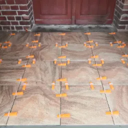 Układanie jasnobrązowych płytek podłogowych z użyciem pomarańczowych klipsów poziomujących, widoczna ceglana ściana i drewniane drzwi.