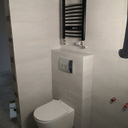 Pan Krzysztof Rogalski - Wykończona łazienka: białe płytki, wisząca toaleta z zabudową, czarny grzejnik drabinkowy. Widoczne detale instalacji i wykończenia.