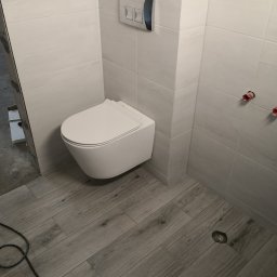 Pan Krzysztof Rogalski - Nowoczesna, biała toaleta podwieszana w wykończonej łazience z płytkami imitującymi drewno na podłodze i jasnymi, kwadratowymi płytkami na ścianach.