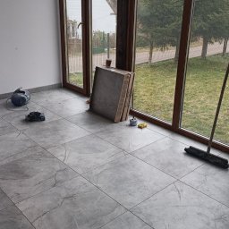 Pan Krzysztof Rogalski - Ułożone szare płytki podłogowe w jasnym pomieszczeniu z dużymi oknami. Widoczne narzędzia glazurnicze i paczki płytek. Naturalne światło dzienne.