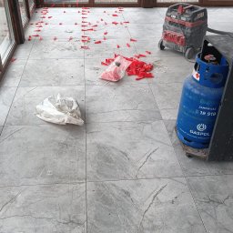 Pan Krzysztof Rogalski - Układanie szarych płytek podłogowych w oranżerii, widoczne czerwone klipsy poziomujące, odkurzacz przemysłowy i butla gazowa. Praca w toku, światło dzienne.