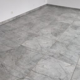 Pan Krzysztof Rogalski - Szare, marmurkowe płytki podłogowe ułożone w pustym pomieszczeniu z białymi ścianami i gniazdkami elektrycznymi przy podłodze. Widok z góry, neutralne oświetlenie.
