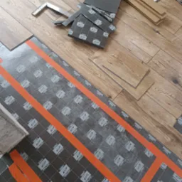 Układanie paneli podłogowych na podkładzie z folią izolacyjną z pomarańczowymi liniami. Widoczne narzędzia i fragmenty paneli. Ujęcie z g&oacute;ry, w trakcie montażu.