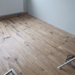 Pan Krzysztof Rogalski - Ułożona podłoga z paneli drewnopodobnych w jasnym odcieniu, widoczne narzędzia pomiarowe i fragment ściany z grzejnikiem. Układanie paneli podłogowych w trakcie prac.