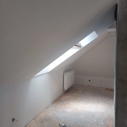 Pan Krzysztof Rogalski - Poddasze po malowaniu na biało, z oknem dachowym i grzejnikiem. Na podłodze widoczne narzędzia malarskie i puszka farby. Ściany gładkie, świeżo pomalowane.