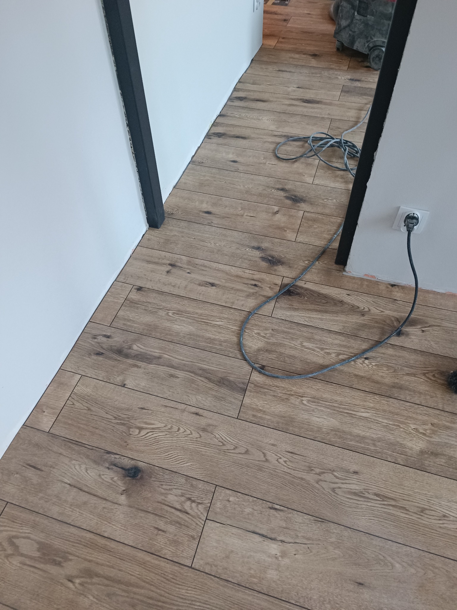 Ułożona podłoga z paneli drewnopodobnych, widoczny fragment ściany z gniazdkiem i kabel zasilający. W tle odkurzacz przemysłowy. Detal wykończenia wnętrza.
