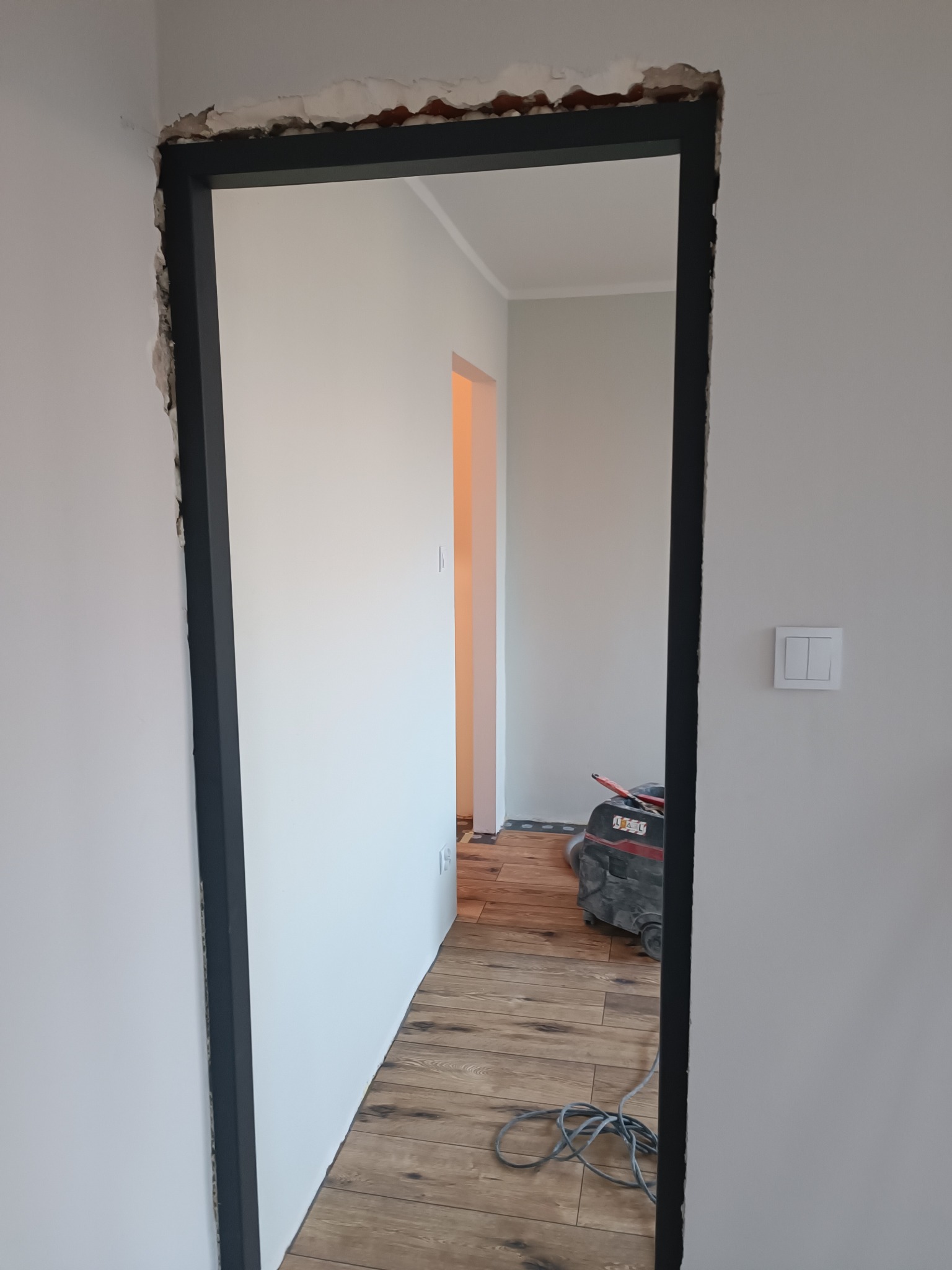 Montaż ościeżnicy w surowym otworze drzwiowym na tle drewnopodobnej podłogi i maszyny do szlifowania. Widoczne niedoskonałości ściany.