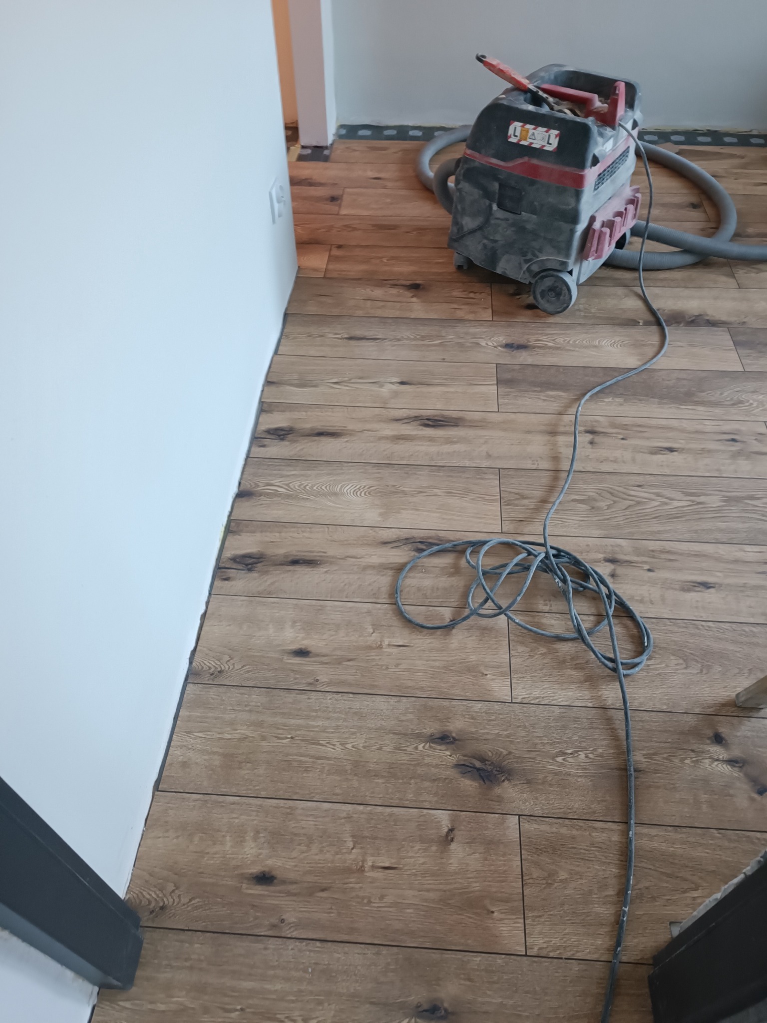 Ułożona drewnopodobna podłoga w jasnym odcieniu brązu, odkurzacz przemysłowy przy ścianie. Widoczne listwy przypodłogowe i kabel od odkurzacza na podłodze.