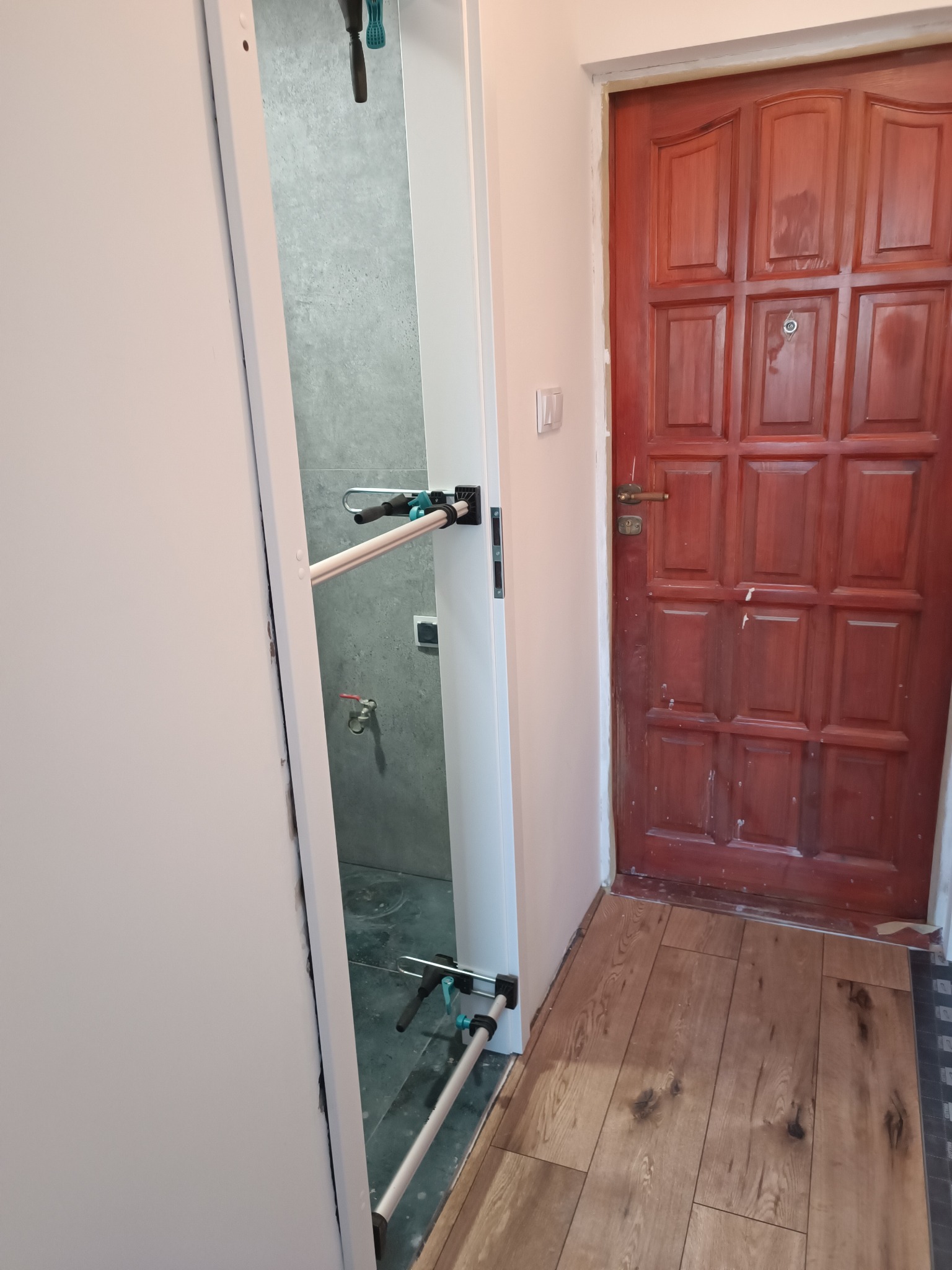 Otwarta biała rama drzwi ukazuje szarą łazienkę z narzędziami. Obok, drewniane panele podłogowe i czerwone drzwi wejściowe.