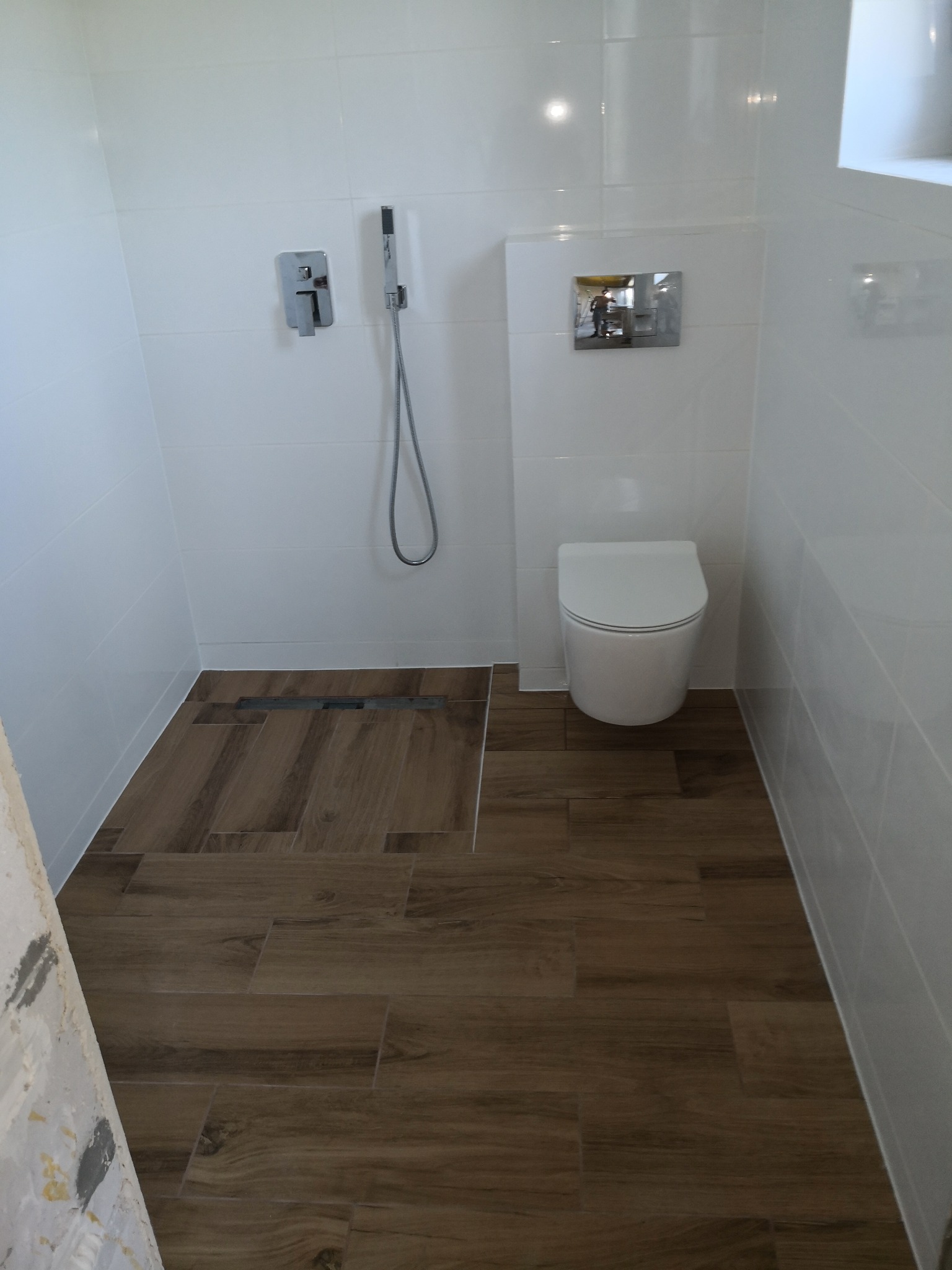 Nowoczesna łazienka po remoncie: białe płytki ścienne, drewnopodobna podłoga z odpływem liniowym, wisząca toaleta i minimalistyczna armatura.