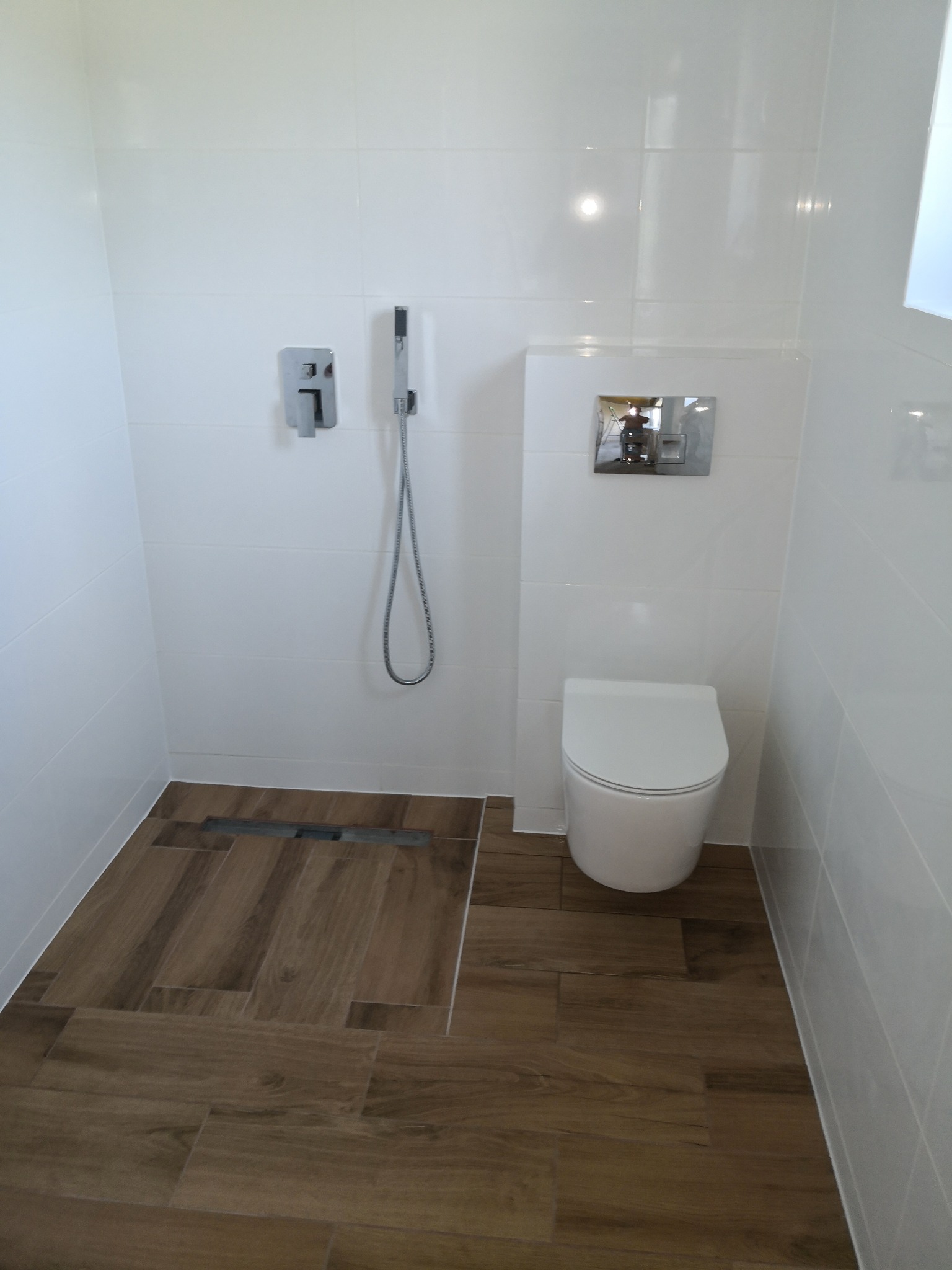 Nowoczesna łazienka z białymi płytkami ściennymi i drewnopodobną podłogą. Uwagę zwraca odpływ liniowy w strefie prysznicowej i minimalistyczna armatura.