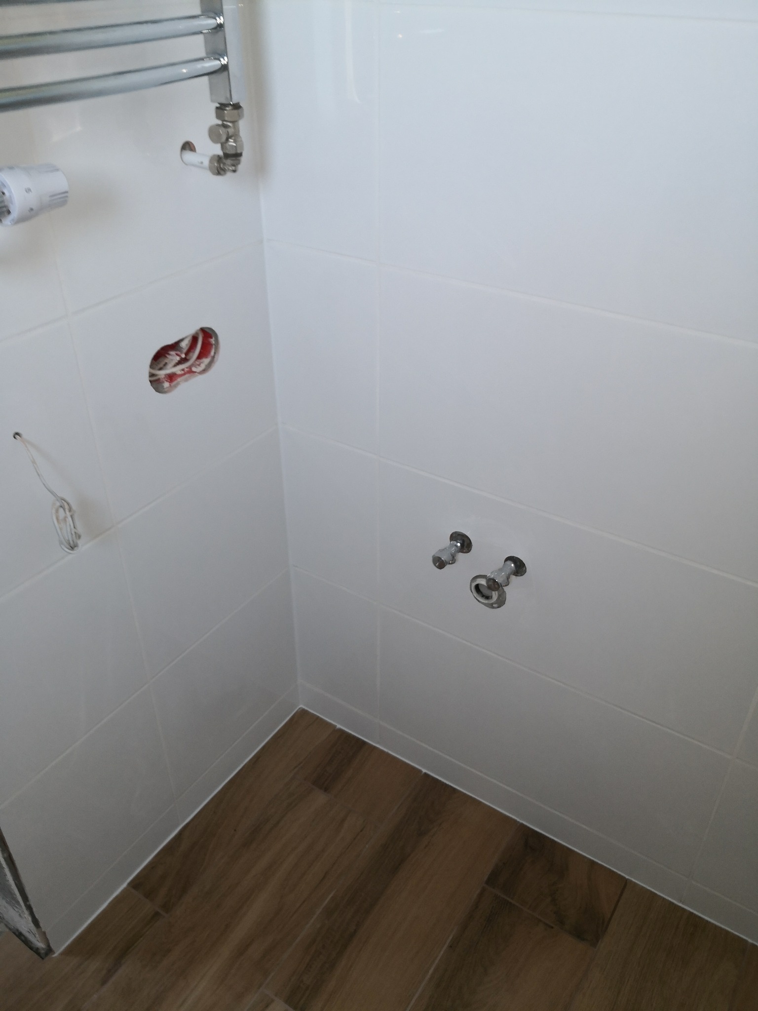 Wnętrze łazienki w trakcie prac wykończeniowych: białe płytki ścienne, imitacja drewna na podłodze, widoczne instalacje i otwór na przewody.