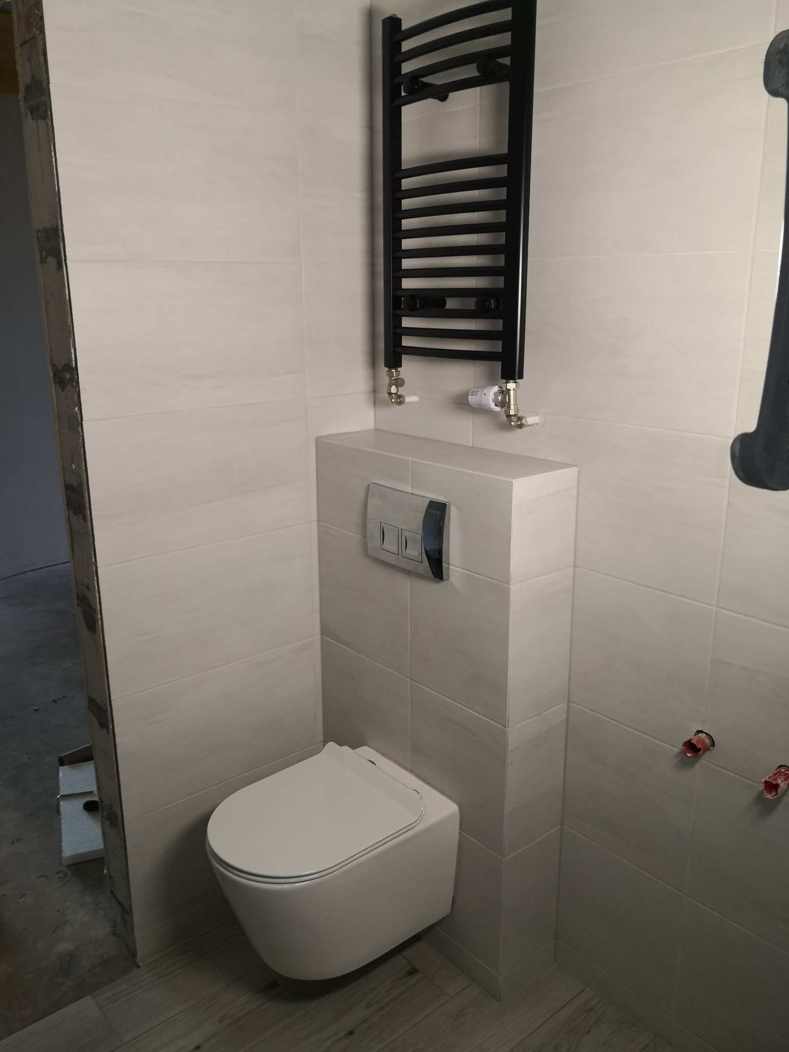 Wykończona łazienka: białe płytki, wisząca toaleta z zabudową, czarny grzejnik drabinkowy. Widoczne detale instalacji i wykończenia.