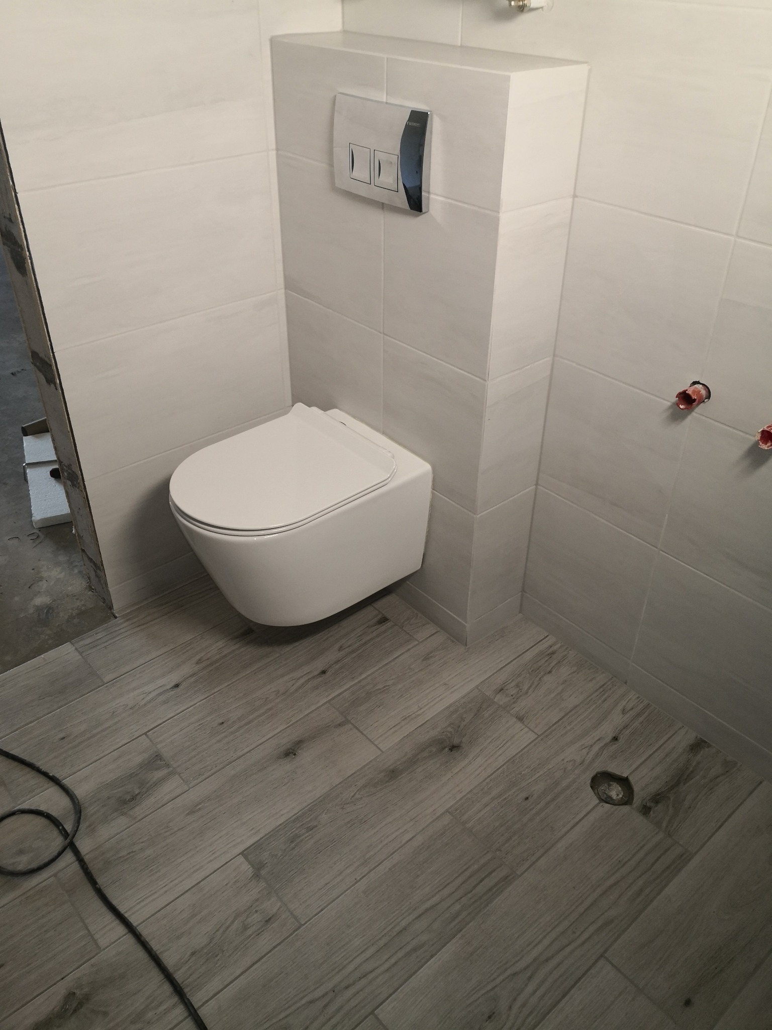 Nowoczesna, biała toaleta podwieszana w wykończonej łazience z płytkami imitującymi drewno na podłodze i jasnymi, kwadratowymi płytkami na ścianach.