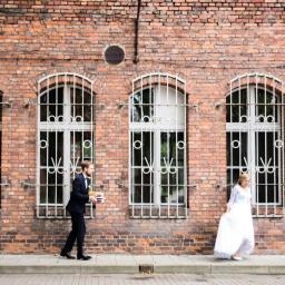 Niebieskie Migdały Zosia Gnat - Para młoda w plenerze: pan młody w garniturze niesie książki, panna młoda w białej sukni spaceruje przed ceglaną ścianą z oknami z kratami.