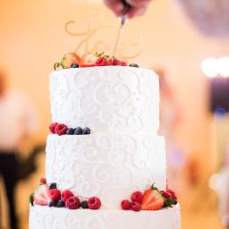 Niebieskie Migdały Zosia Gnat - Trzypiętrowy tort weselny z białym kremem i dekoracją z truskawek, malin i borówek, krojony nożem przez osobę z niewyraźnym tłem.