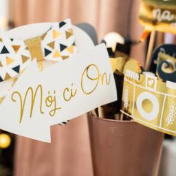 Niebieskie Migdały Zosia Gnat - Rekwizyty do fotobudki: biała strzałka z napisem 'Mój ci On', muszka w złote trójkąty, złota kamera i wąsy, stojące w miedzianym wiaderku.