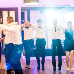 Niebieskie Migdały Zosia Gnat - Grupa młodych ludzi w formalnych strojach tańczy trzymając się za ramiona na parkiecie oświetlonym punktowymi światłami podczas imprezy.