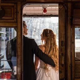 Niebieskie Migdały Zosia Gnat - Para młoda w zabytkowym tramwaju, panna młoda w sukni z koronkowym wzorem, pan młody w czarnym garniturze, widok z tyłu, ciepłe światło wpadające przez okno.
