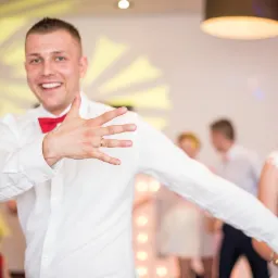 Uśmiechnięty pan młody z czerwoną muchą, wyciągniętą ręką i obrączką na palcu, obok uśmiechnięta panna młoda w białej sukni na tle bawiących się gości.