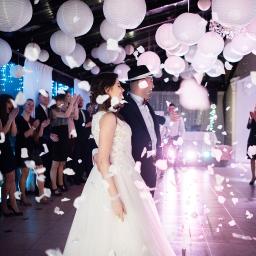 Niebieskie Migdały Zosia Gnat - Para młoda w tańcu, obsypywana płatkami kwiatów, w tle goście klaszczą, dekoracja sali z lampionami, oświetlenie punktowe.