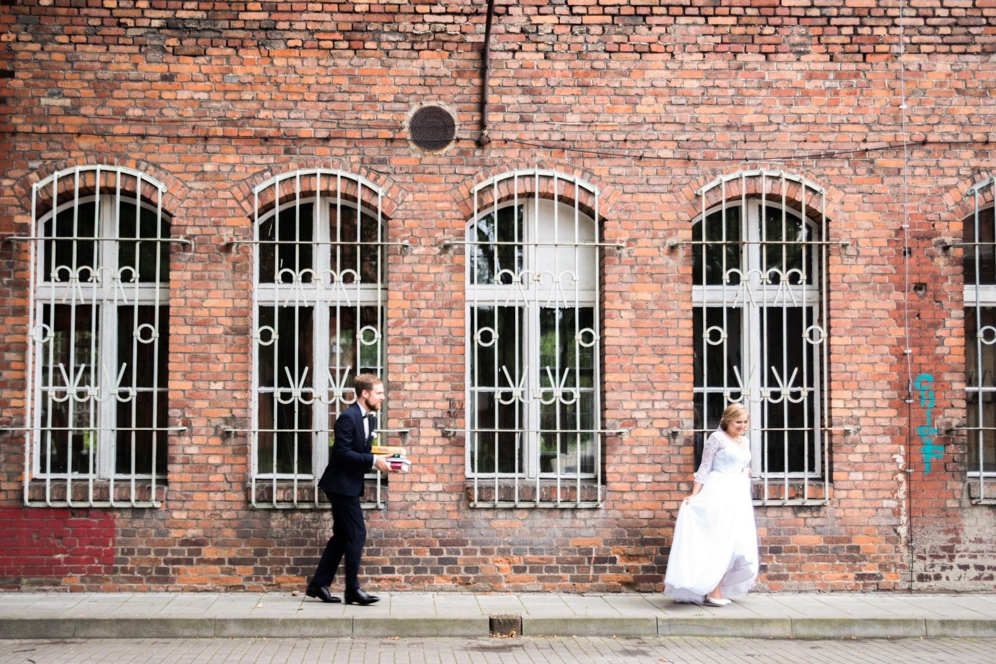 Para młoda w plenerze: pan młody w garniturze niesie książki, panna młoda w białej sukni spaceruje przed ceglaną ścianą z oknami z kratami.