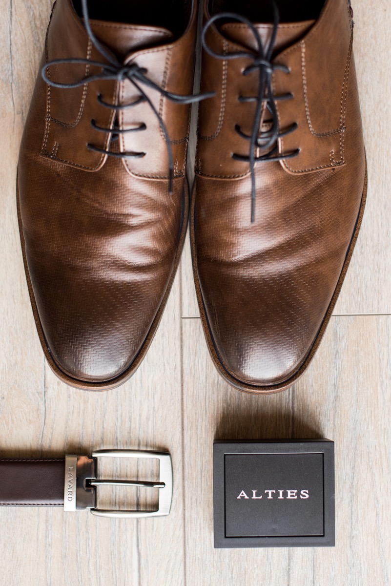 Brązowe skórzane buty z czarnymi sznurówkami, brązowy pasek ze srebrną klamrą marki Lavard i czarne pudełko marki Alties na jasnym drewnianym tle.
