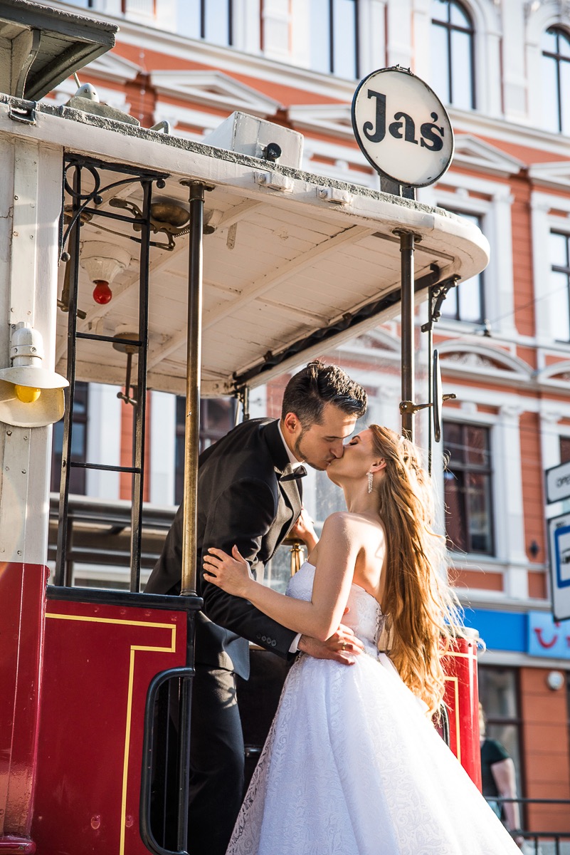 Para młoda w pocałunku wewnątrz zabytkowego tramwaju z napisem 'Jaś' na dachu, na tle miejskiej architektury.