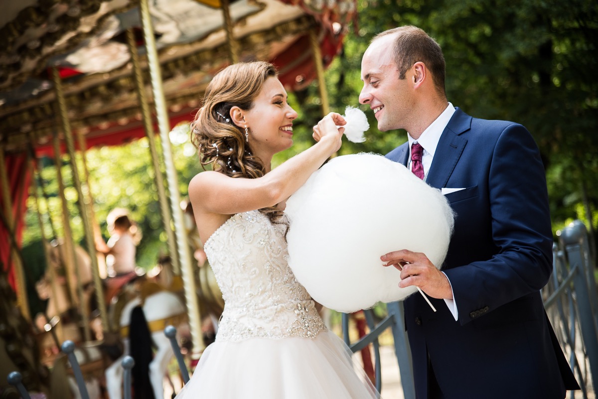 Uśmiechnięta panna młoda w zdobionej sukni częstuje pana młodego watą cukrową na tle karuzeli.
