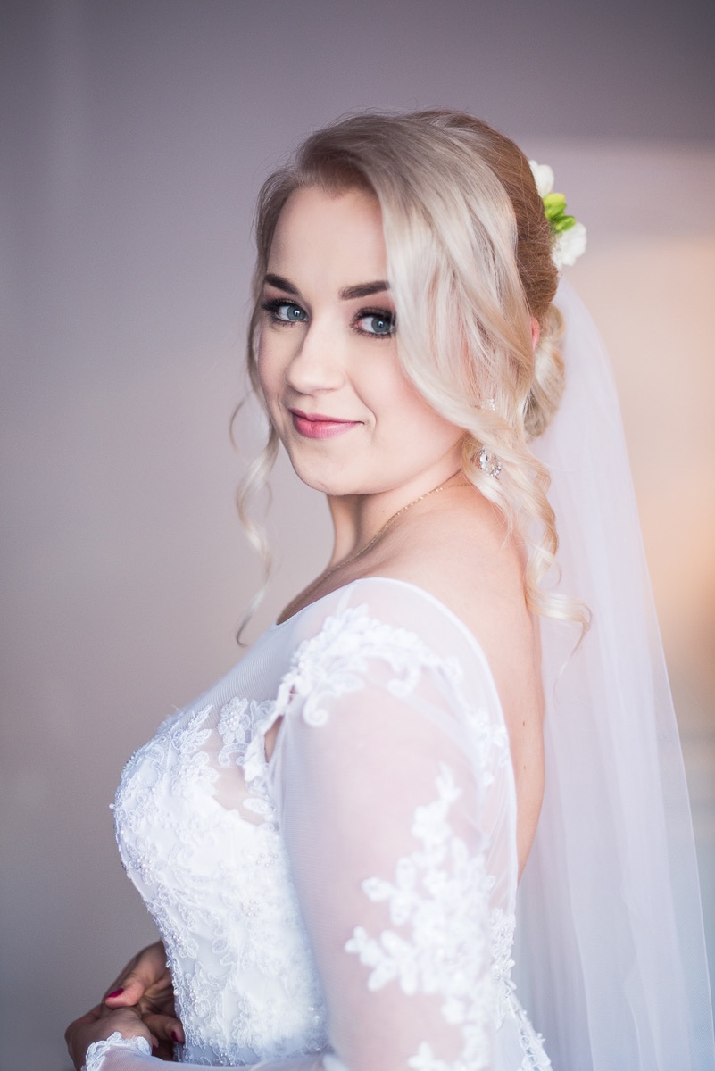 Uśmiechnięta panna młoda w białej koronkowej sukni z tiulowym welonem i kwiatami we włosach, spoglądająca przez ramię.