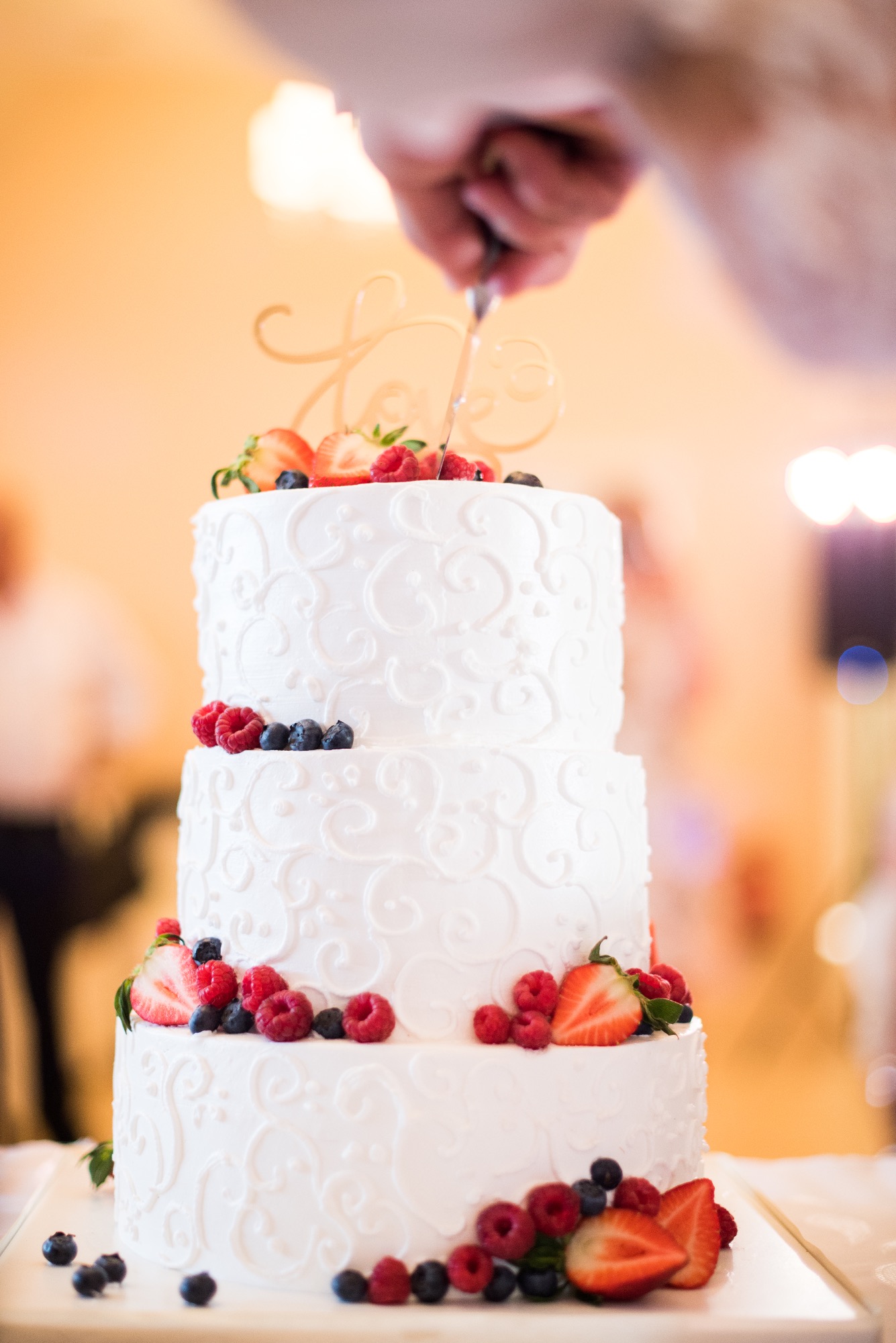 Trzypiętrowy tort weselny z białym kremem i dekoracją z truskawek, malin i borówek, krojony nożem przez osobę z niewyraźnym tłem.