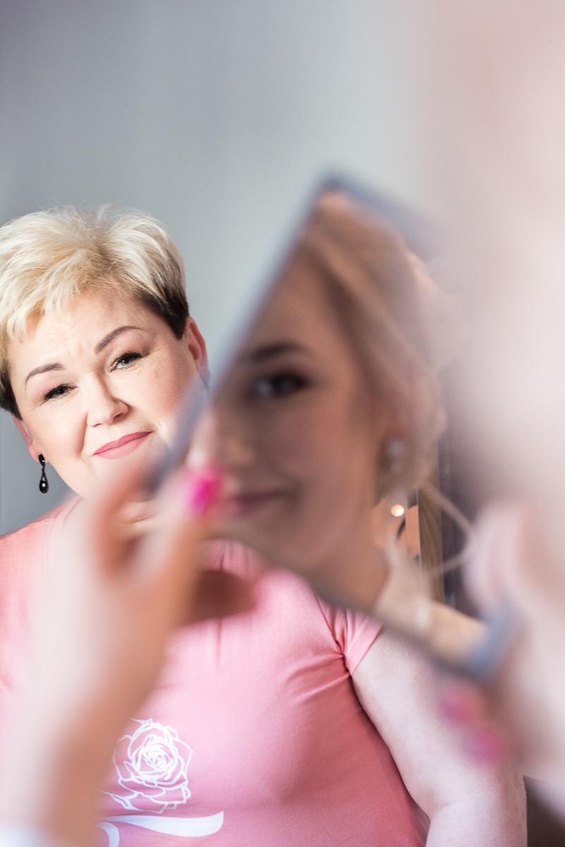 Kobieta w różowej koszulce z motywem róży spogląda z uśmiechem na odbicie w lustrze, gdzie widać fragment twarzy innej kobiety z delikatnym makijażem.