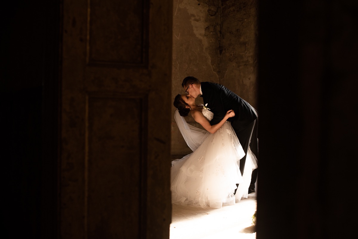 Para młoda całuje się w zabytkowym wnętrzu, widoczni przez framugę drzwi, on w czarnym garniturze, ona w białej sukni z welonem, oświetleni promieniami słońca wpadającymi przez okno.