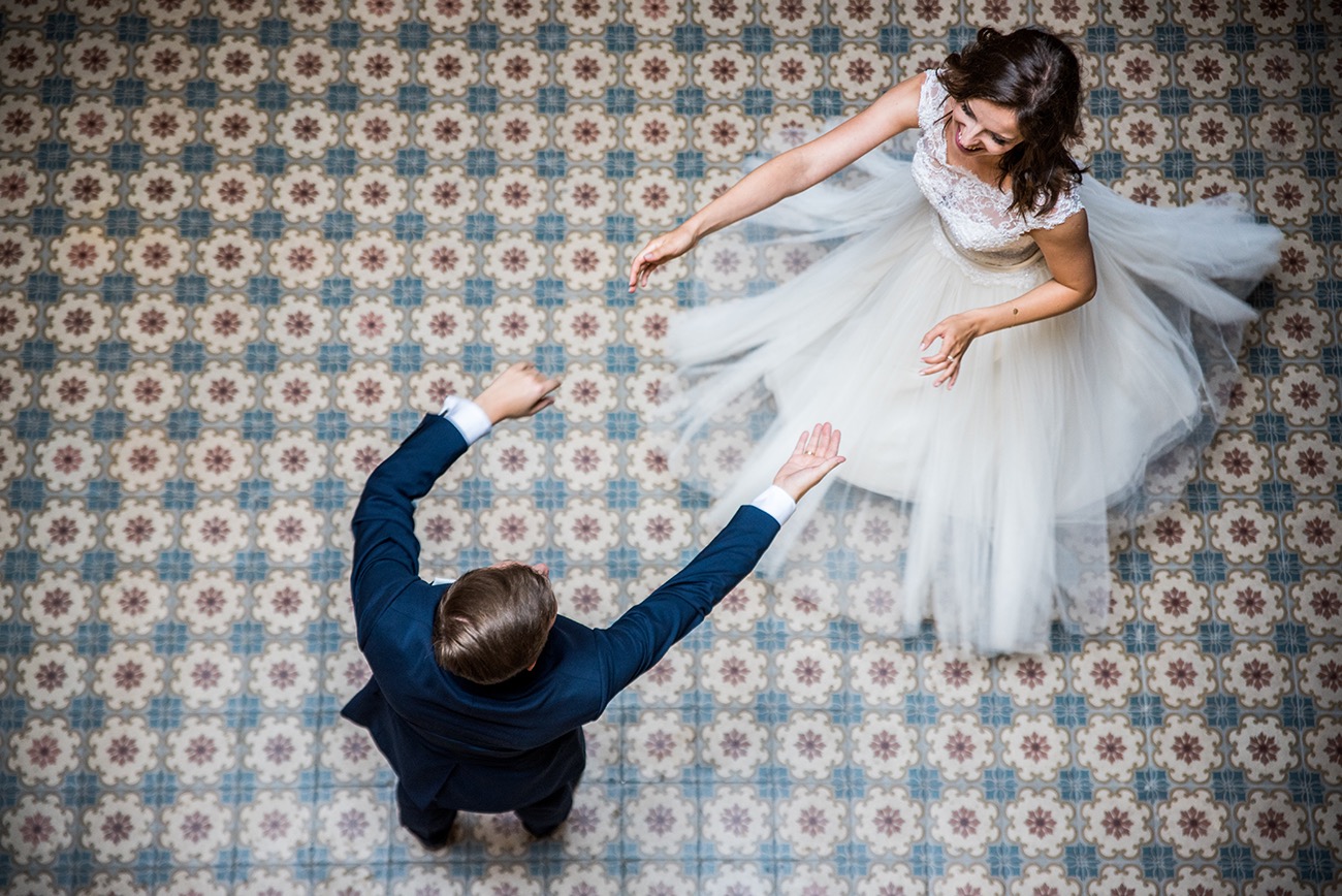 Widok z góry na parę młodą w tańcu na wzorzystej podłodze, panna młoda w koronkowej sukni z tiulowym dołem, pan młody w granatowym garniturze, podniesione ręce w geście radości.