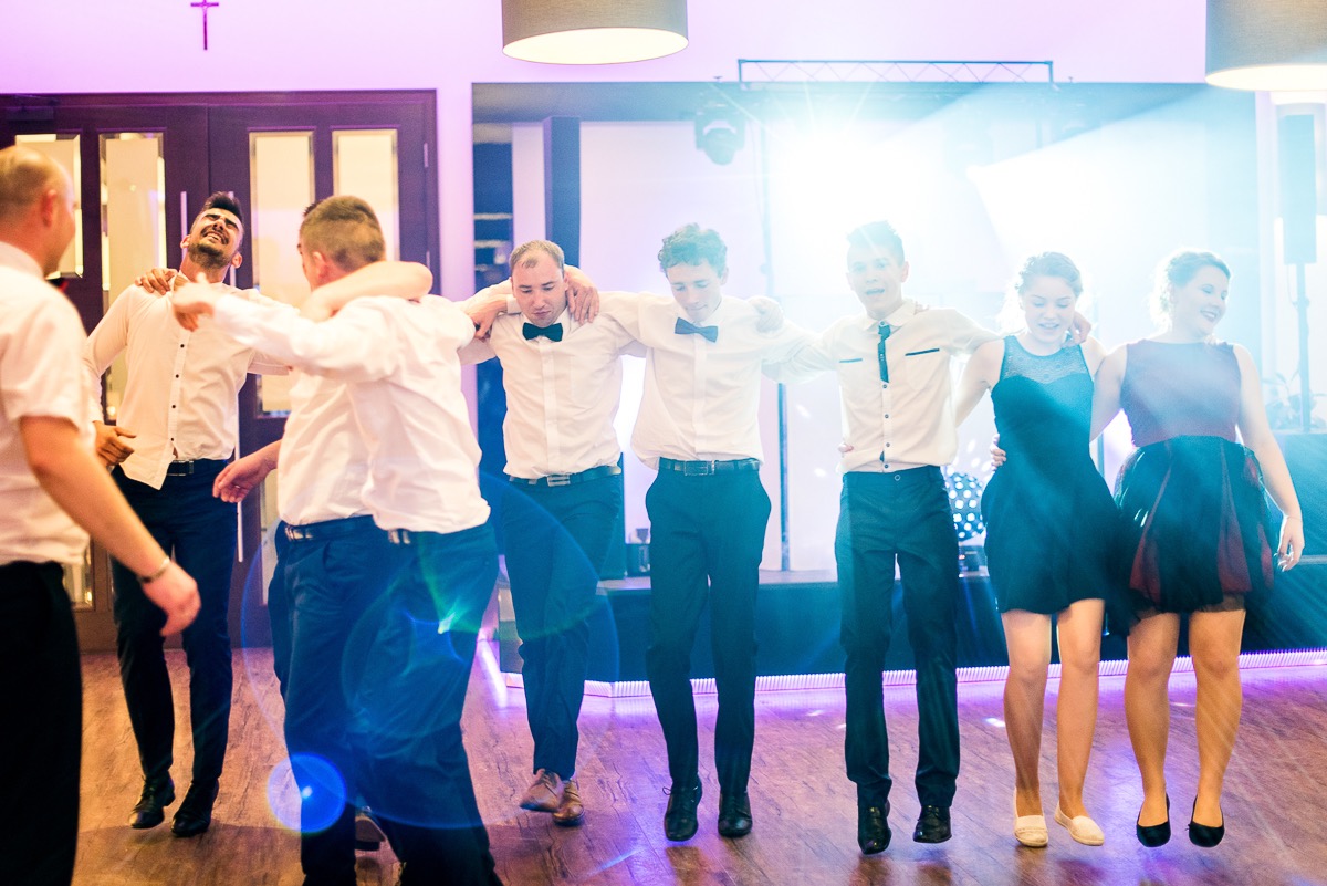 Grupa młodych ludzi w formalnych strojach tańczy trzymając się za ramiona na parkiecie oświetlonym punktowymi światłami podczas imprezy.