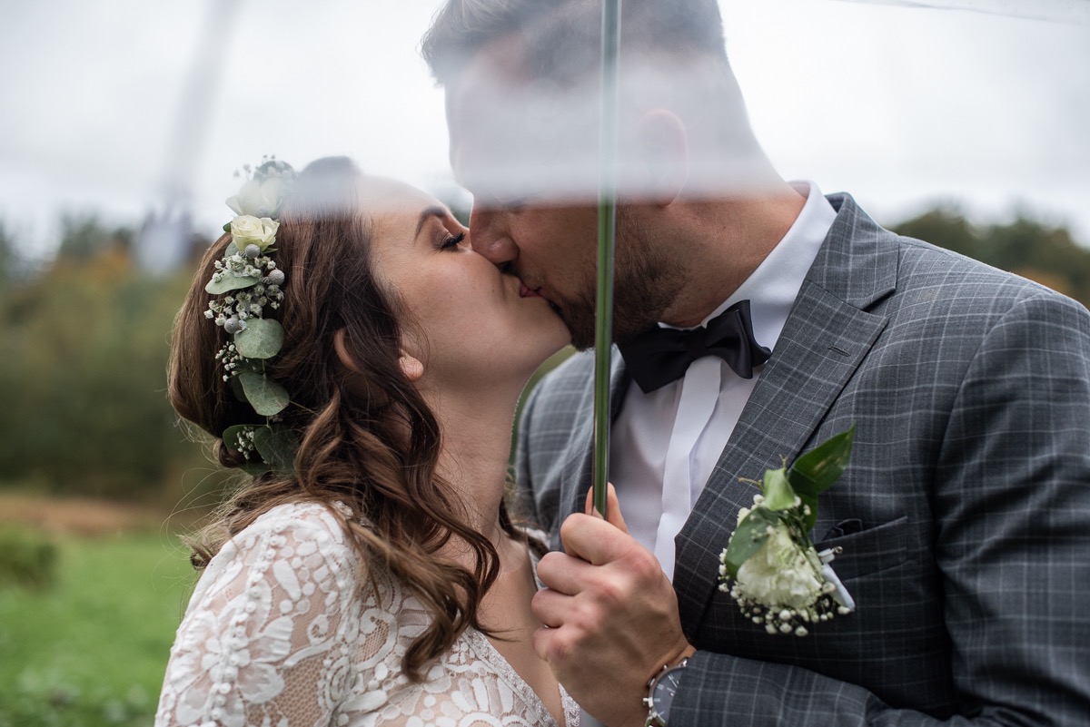 Pocałunek nowożeńców pod parasolem: panna młoda w koronkowej sukni i wianku, pan młody w szarym garniturze w kratę z muchą, zbliżenie na twarze.