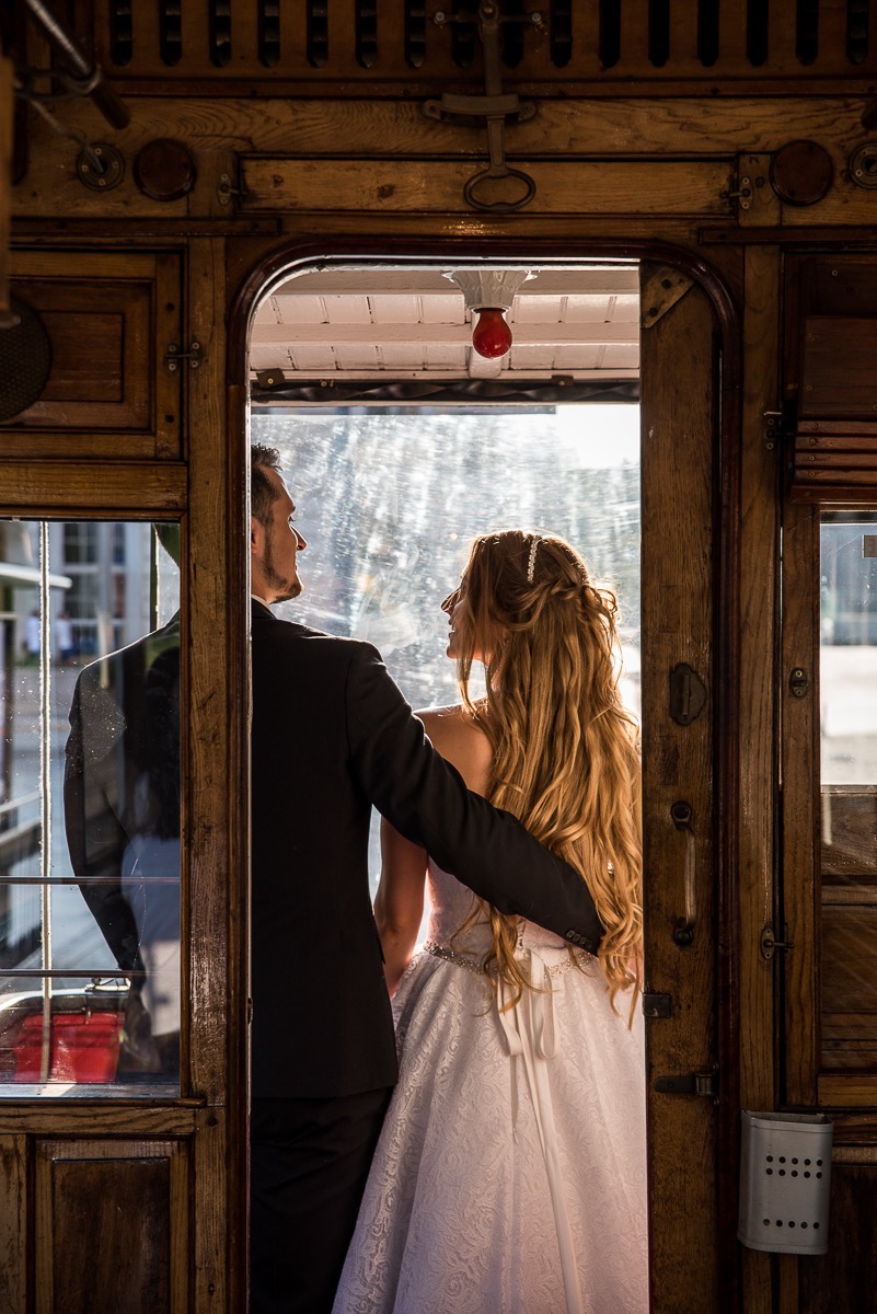 Para młoda w zabytkowym tramwaju, panna młoda w sukni z koronkowym wzorem, pan młody w czarnym garniturze, widok z tyłu, ciepłe światło wpadające przez okno.