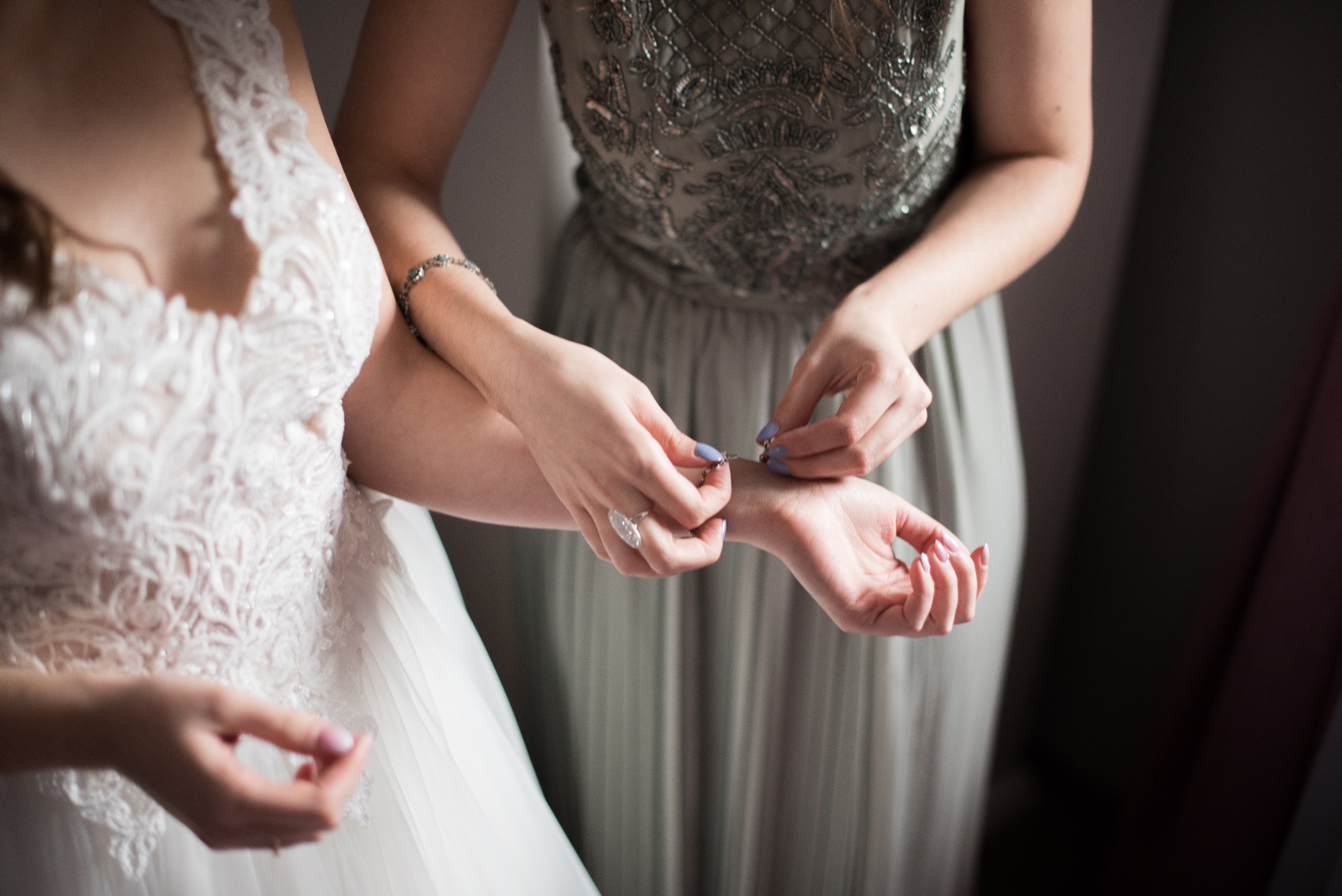Panna młoda w białej sukni z koronkowym wykończeniem, pomagająca druhnie zapiąć srebrną bransoletkę na nadgarstku. Druhna ma na sobie szarą sukienkę z ozdobnymi aplikacjami, a na palcu duży...