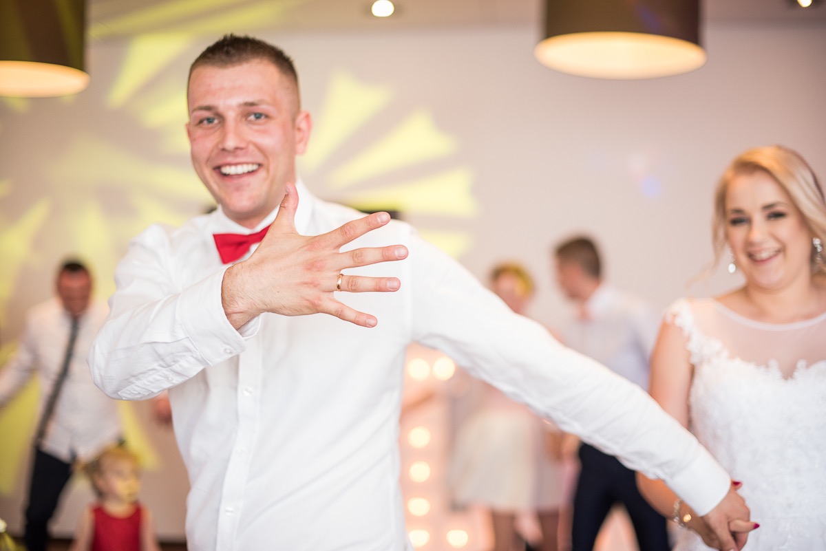 Uśmiechnięty pan młody z czerwoną muchą, wyciągniętą ręką i obrączką na palcu, obok uśmiechnięta panna młoda w białej sukni na tle bawiących się gości.