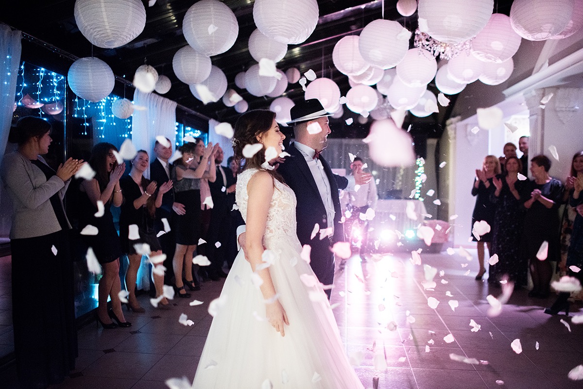 Para młoda w tańcu, obsypywana płatkami kwiatów, w tle goście klaszczą, dekoracja sali z lampionami, oświetlenie punktowe.
