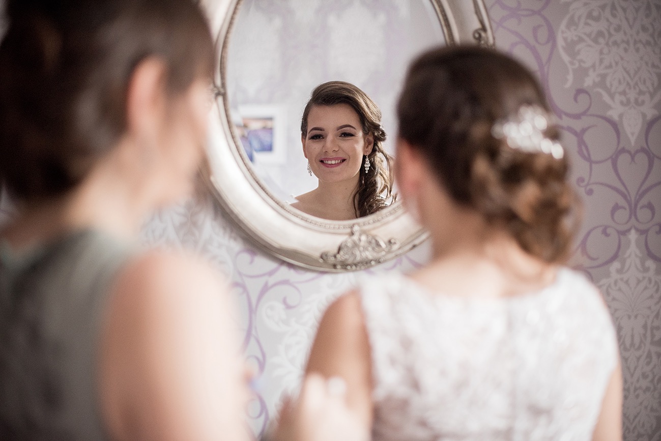 Uśmiechnięta kobieta z upiętymi włosami i kolczykami, widoczna w ozdobnym lustrze podczas przygotowań, z dwiema osobami w tle.