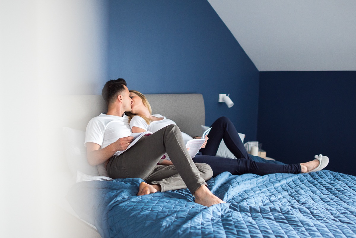 Para całująca się leżąc na łóżku z niebieską narzutą, czytając książki w pokoju z granatową ścianą.