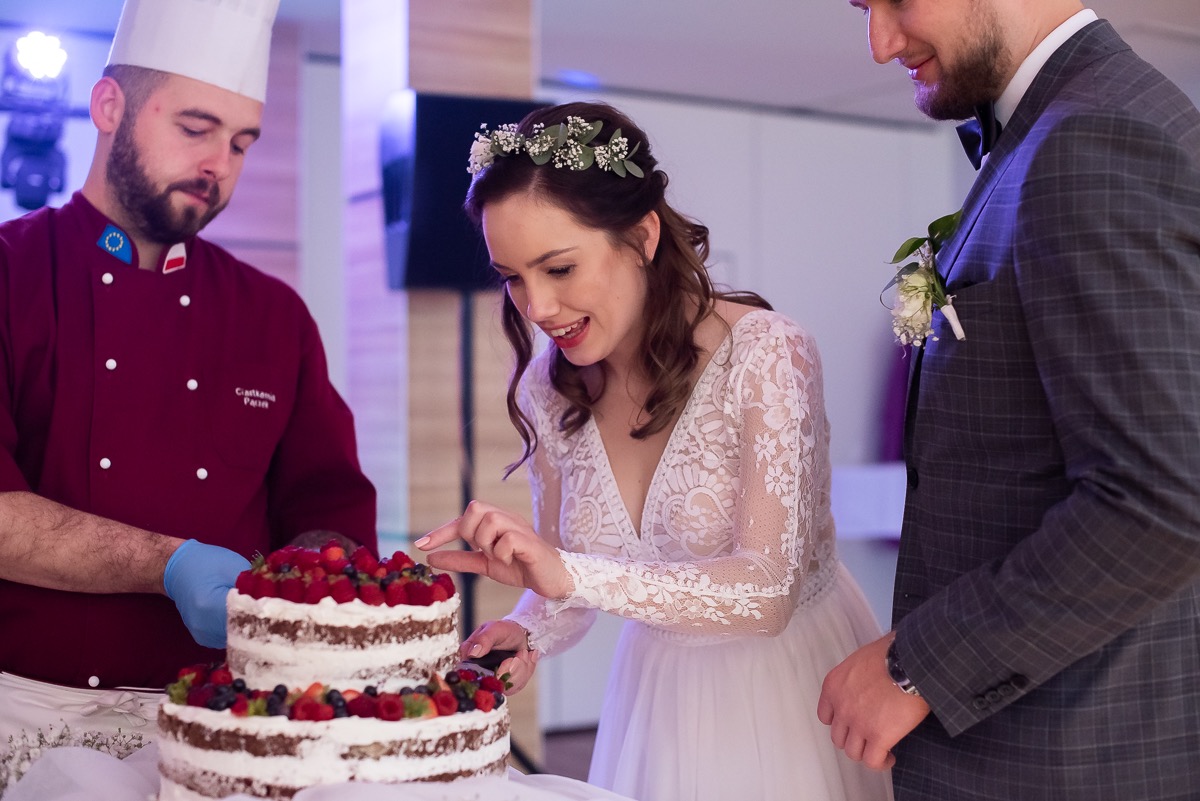 Panna młoda w koronkowej sukni i wianku kroi dwupiętrowy tort udekorowany świeżymi owocami, obok pan młody w garniturze w kratę i kucharz w uniformie z rękawicami.