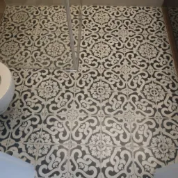 Czarno-biała, wzorzysta podłoga wyłożona płytkami w łazience, widoczny fragment toalety i kabiny prysznicowej z metalowymi elementami.