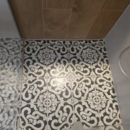 Fragment wykończonej łazienki z dekoracyjnymi, czarno-białymi płytkami podłogowymi i drewnopodobną okładziną sufitową, widoczny fragment kabiny prysznicowej i grzejnika drabinkowego.