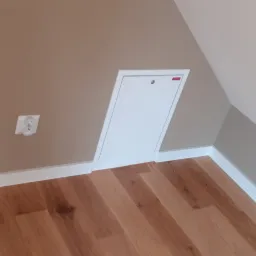 Poddasze po remoncie: drewnopodobne panele podłogowe, beżowa ściana z gniazdkiem w kształcie misia i białe listwy przypodłogowe oraz białe drzwiczki do schowka na poddaszu.