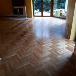 Parkiet ułożony w jodełkę, widok na salon z dużym oknem i kominkiem, ślady po cyklinowaniu widoczne na powierzchni.