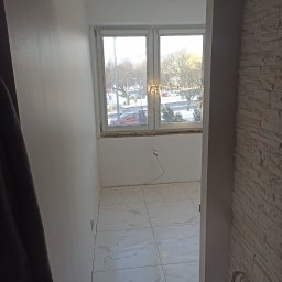 NIKO - Wykończony balkon z białymi płytkami podłogowymi i ścianą z dekoracyjnej cegły. Widoczne okno z widokiem na ulicę. Ściany pomalowane na biało.