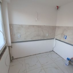 NIKO - Wykończona podłoga z płytek imitujących marmur i częściowo położone płytki ścienne w kuchni. Widoczne puszki elektryczne i listwa montażowa. W tle okno.